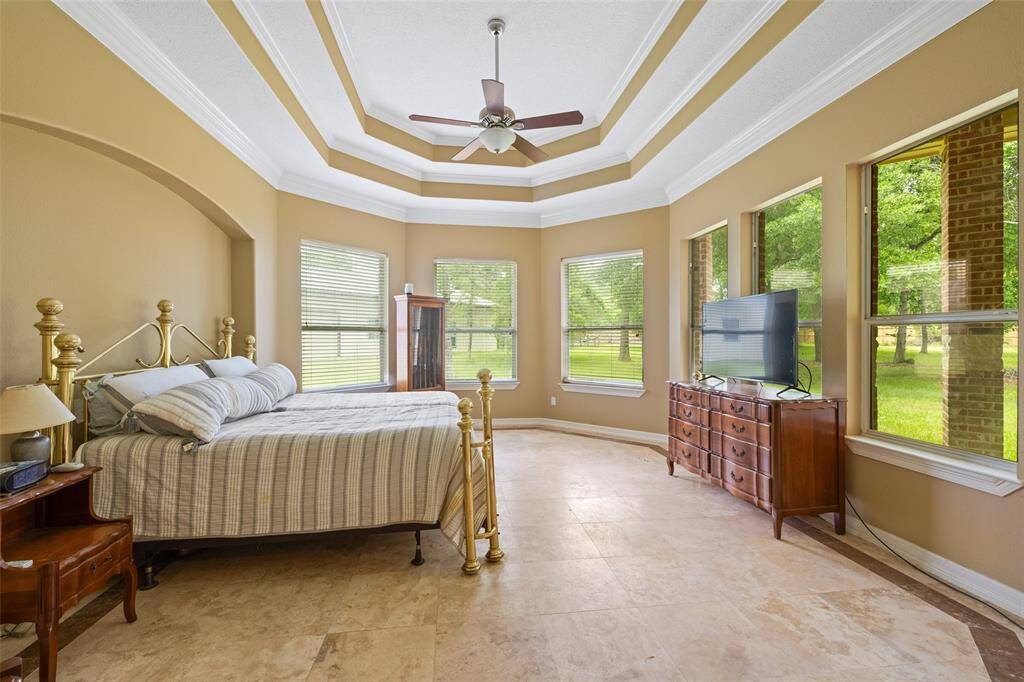 This spacious bedroom features a tray ceiling with a fan, large windows offering plenty of natural light and views of the outside, tile flooring, creating a bright and inviting atmosphere.