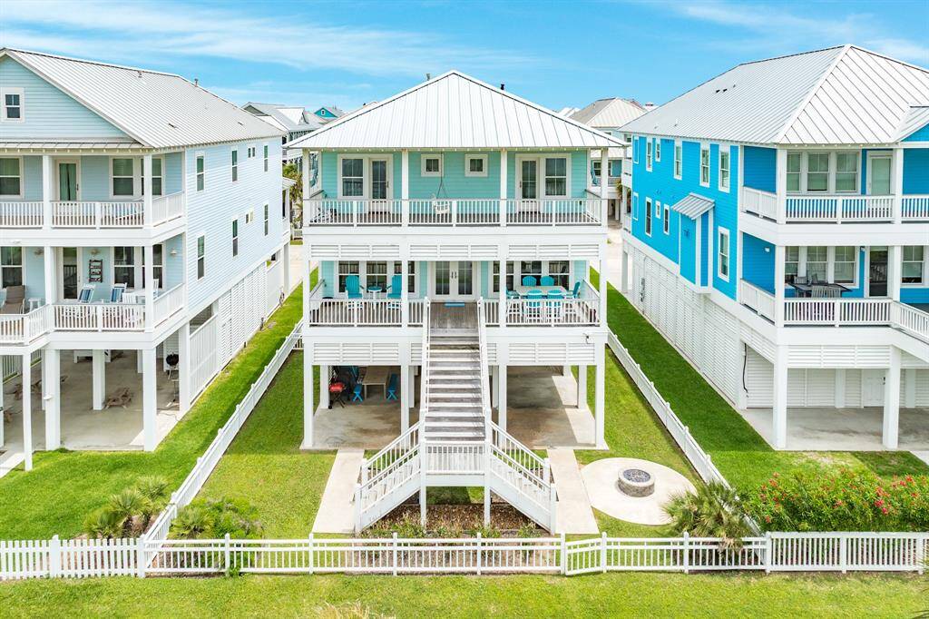 3810 Abalone a charming coastal home elevated on stilts, featuring a spacious two-story design with large balconies and a metal roof. The property is flanked by similar homes and includes a fenced yard with a fire pit, offering a blend of modern amenities and beachside living.