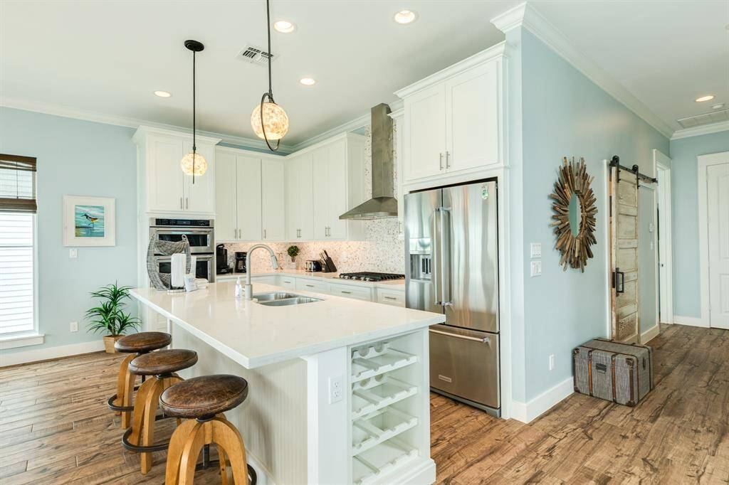 This bright, modern kitchen features a large island with seating, stainless steel appliances, white cabinetry, and stylish pendant lighting. The rustic flooring and sliding barn door from the 1900's adds a cozy charm.