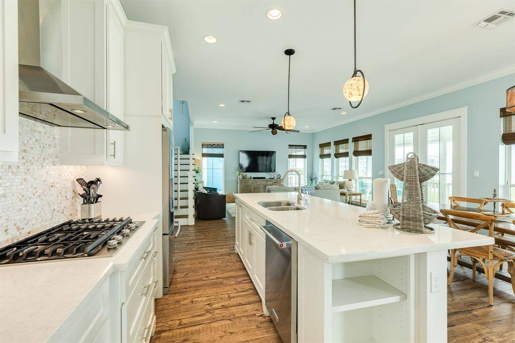 The kitchen features sleek white cabinetry, a gas stove, and a large island with a sink. The space is illuminated by pendant lighting and natural light from large windows. It flows seamlessly into a cozy living room with wood flooring and a ceiling fan, offering a bright and airy atmosphere.