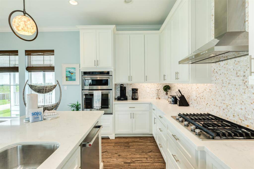 This modern kitchen features sleek white cabinetry, a gas stovetop, and stainless steel finish appliances. The space is bright and airy, with a large Cambria island, elegant pendant lighting, and a stylish mosaic backsplash.