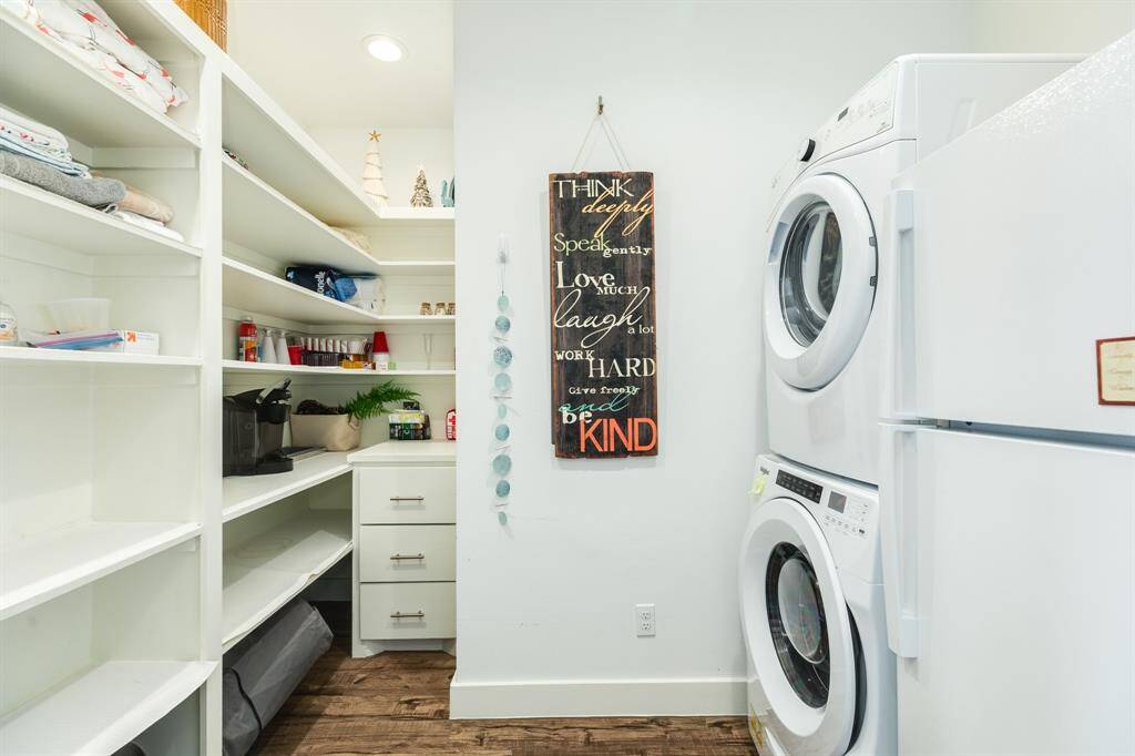 Home features a well-organized utility room with white shelves and cabinets for storage, a stacked washer and dryer, and a wall-mounted inspirational sign. The room has a bright, clean look with wood-style flooring.