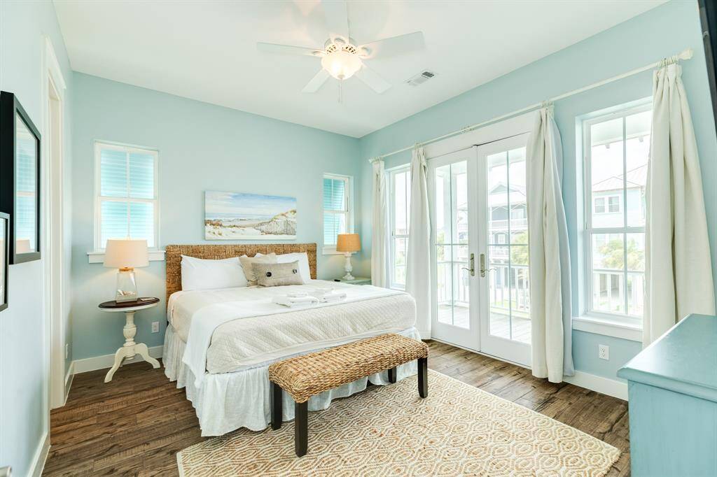 Bright and airy bedroom 1 with light blue walls, featuring a cozy bed, wicker accents, and large windows leading to a balcony. The room has a ceiling fan, wood flooring, and a serene, coastal decor.
