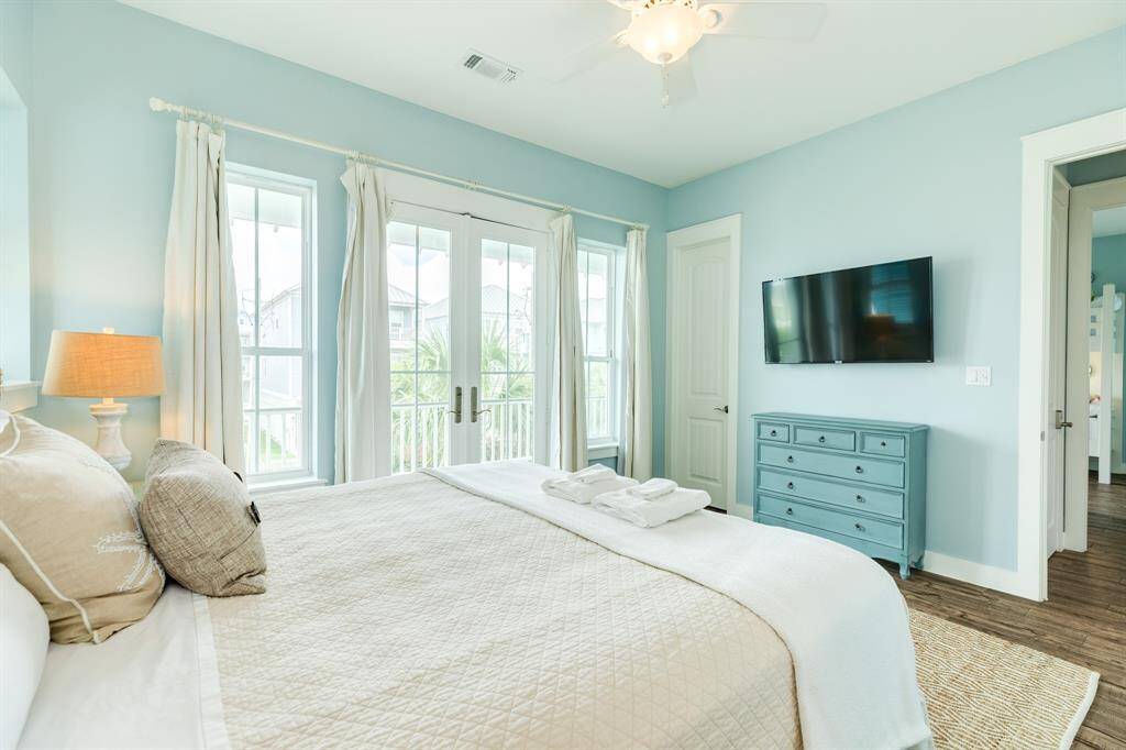 Bright and airy bedroom 1 with soft blue walls, featuring a large bed, ceiling fan, and wall-mounted TV. It includes a dresser, double doors with curtains leading to the 1st floor balcony, and ample natural light from multiple windows.