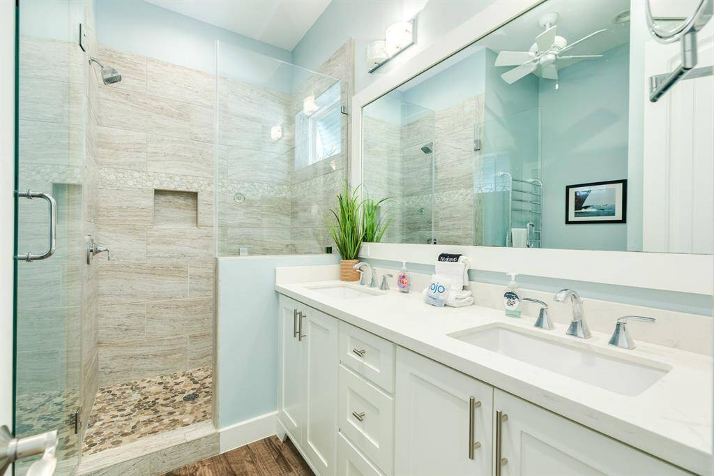 This modern ensuite bathroom features a spacious glass-enclosed shower with stylish tilework and pebble flooring. It includes a large double vanity with sleek fixtures, ample storage, and a bright, airy feel enhanced by a large mirror and soft lighting.