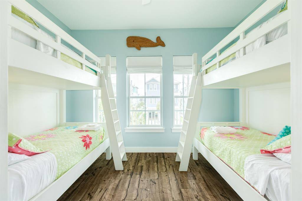 Bright and airy bunk room featuring two sets of white bunk beds with colorful bedding, light blue walls, and wooden floors. Large windows provide natural light, and a playful whale decoration adds charm.