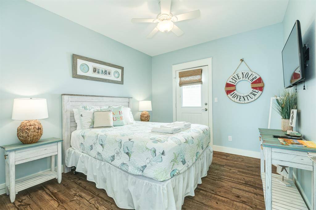 Bedroom 3 features a double bed with a coastal-themed bedspread, complemented by light blue walls and hardwood floors. It includes rustic nightstands with lamps, a wall-mounted TV, and a decorative "Live Your Adventure" sign, creating a tranquil retreat.