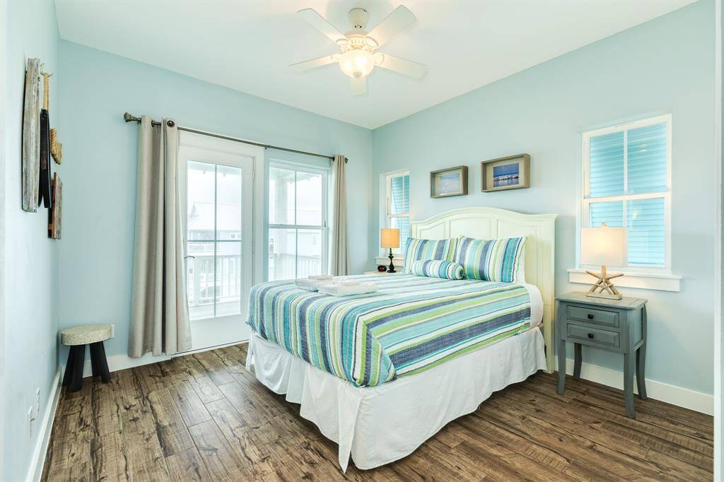 Bright and inviting bedroom 4 with a coastal theme, featuring light blue walls, wood flooring, and a comfortable bed with striped bedding. The space includes a ceiling fan, ample natural light from large windows, and access to the deck through glass doors.