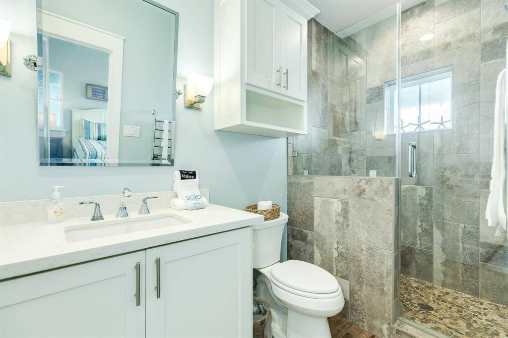 Bath 3 features a modern design with a glass-enclosed shower showcasing stone tilework. It includes a white vanity with a marble countertop, under-sink storage, and a large wall mirror. The room is bright and welcoming, enhanced by soft lighting and neutral tones.