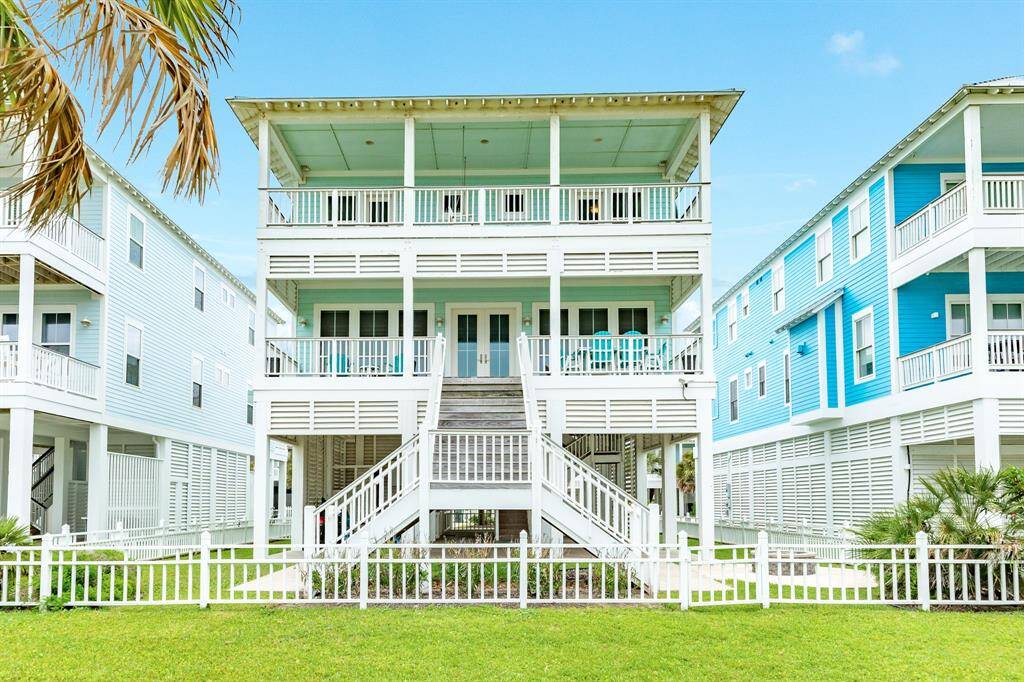 This photo features a charming, elevated coastal-style home with a bright exterior and multiple decks. It has a welcoming staircase, fenced yard, and is flanked by similar vibrant houses, creating a beachside community atmosphere.