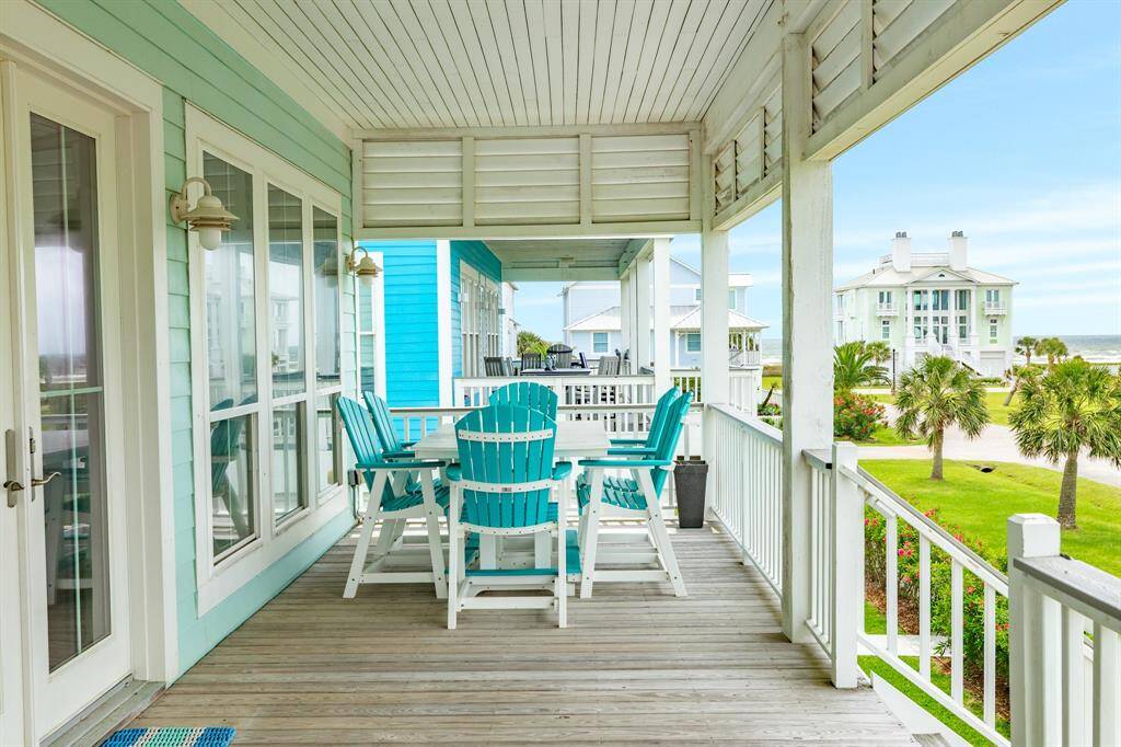This inviting porch features turquoise Adirondack chairs around a table, set against a backdrop of coastal homes and palm trees, offering a relaxing ocean view.