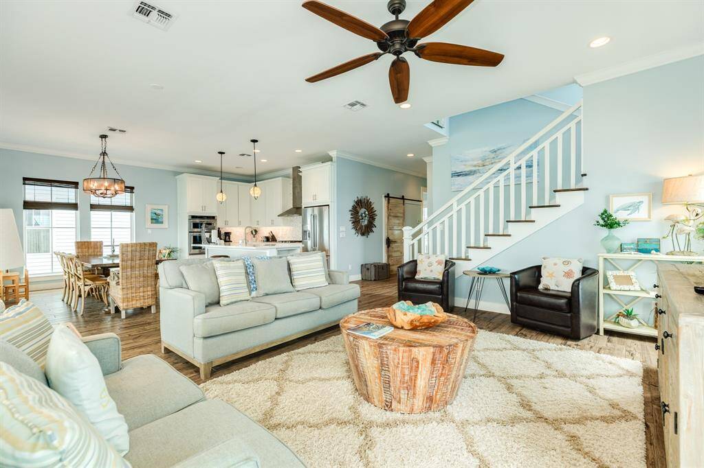 This inviting open-concept living space features a spacious seating area with neutral tones, a rustic wooden coffee table, and a plush rug. The adjacent kitchen boasts modern pendant lighting, sleek appliances, and a dining area with wicker chairs. A stylish staircase adds a touch of elegance, and the overall ambiance is bright and welcoming.