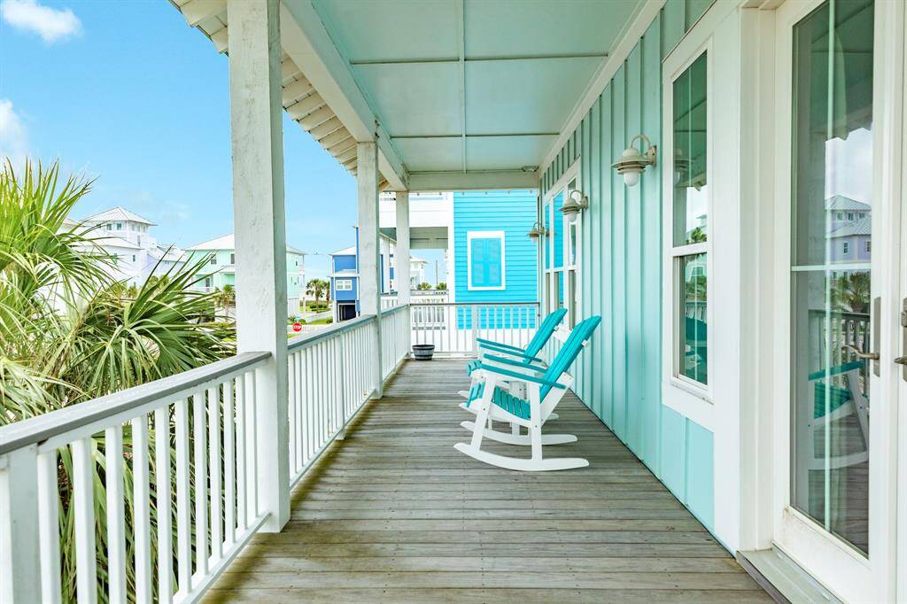 A charming seaside porch with turquoise accents, featuring two rocking chairs and a scenic view of palm trees and neighboring beach houses. Perfect for relaxing and enjoying coastal living.