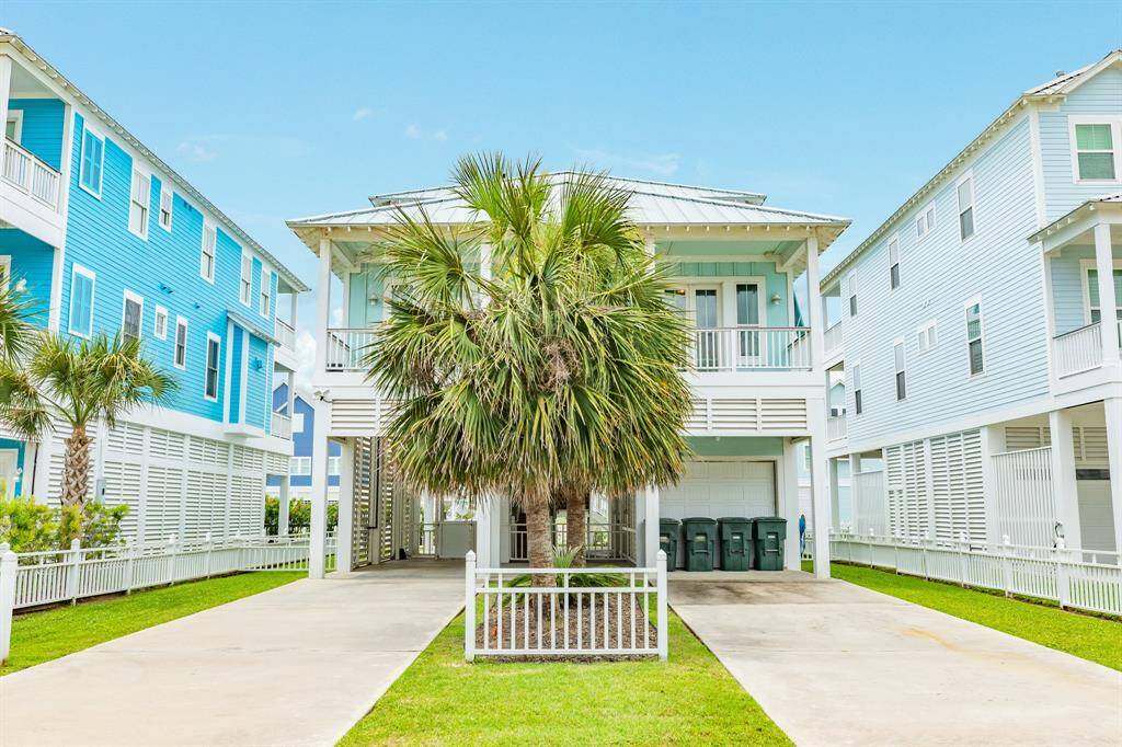 This photo shows a charming coastal-style home with elevated design, featuring a double wide driveway and lush landscaping with palm trees. The house has a light blue exterior, a spacious balcony, and is situated between similar neighboring homes, offering a beachy, community feel.