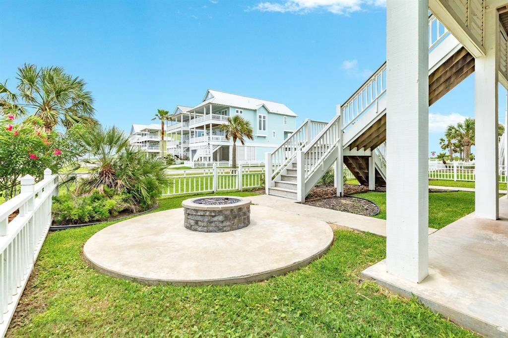 This photo features a charming outdoor space with a circular patio and fire pit, surrounded by lush greenery and white fencing. A staircase leads to an elevated deck for coastal living.