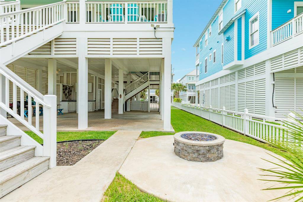 This photo showcases a coastal-style home with elevated construction, featuring spacious covered entertainment area and an outdoor fire pit. The house is surrounded by a neatly maintained lawn and vibrant blue accents, offering a charming and inviting atmosphere.
