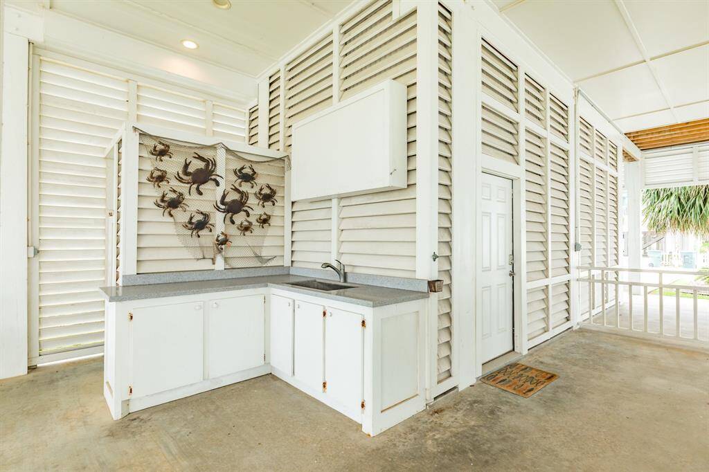 Ground floor entertainment area with white cabinetry and countertops, featuring decorative crab art on the wall and TV Box. The space is bright and airy, with louvered panels and a concrete floor, offering a coastal vibe ideal for entertaining.