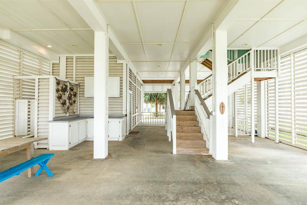 This photo shows an open, airy ground level space with concrete flooring, featuring a staircase leading upstairs. The area has white walls with horizontal slats, providing ventilation and a beachy feel. There’s a built-in counter with cabinets and a decorative wall display, ideal for a coastal lifestyle.