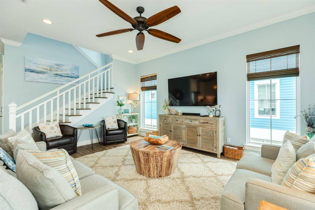 Bright, inviting living room with a modern ceiling fan, large TV, and relaxed seating. Features an elegant staircase, natural light from large windows, and stylish decor. Ideal for relaxation and entertaining.