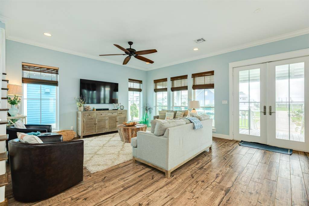 This inviting living room features light blue walls, wood flooring, and ample natural light through large windows. It includes a cozy seating area with a modern sofa and armchairs, a mounted TV, and a ceiling fan. French doors open to an outdoor view, enhancing the airy, relaxed atmosphere.