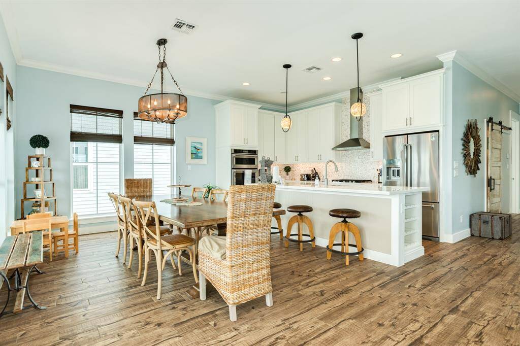 This photo showcases a bright, open-concept kitchen and dining area with a large dining table, and modern light fixtures. The kitchen features white cabinetry, a stainless steel refrigerator, and a central island with bar seating, creating a welcoming space for gatherings.