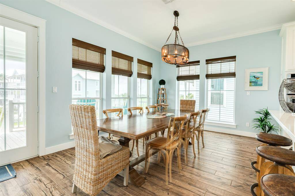 Bright dining area with light blue walls, featuring a large wooden table, wicker and wooden chairs, and a stylish chandelier. The space has abundant natural light from several windows with wooden blinds, offering a cozy and inviting atmosphere.