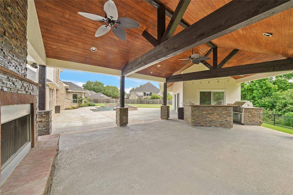 A spacious outdoor patio with a high wooden ceiling, ceiling fans, and a stone fireplace. It features a built-in outdoor kitchen with a grill and ample counter space, perfect for entertaining. The area overlooks the large yard with the pool/spa,  mature trees and neighboring homes.