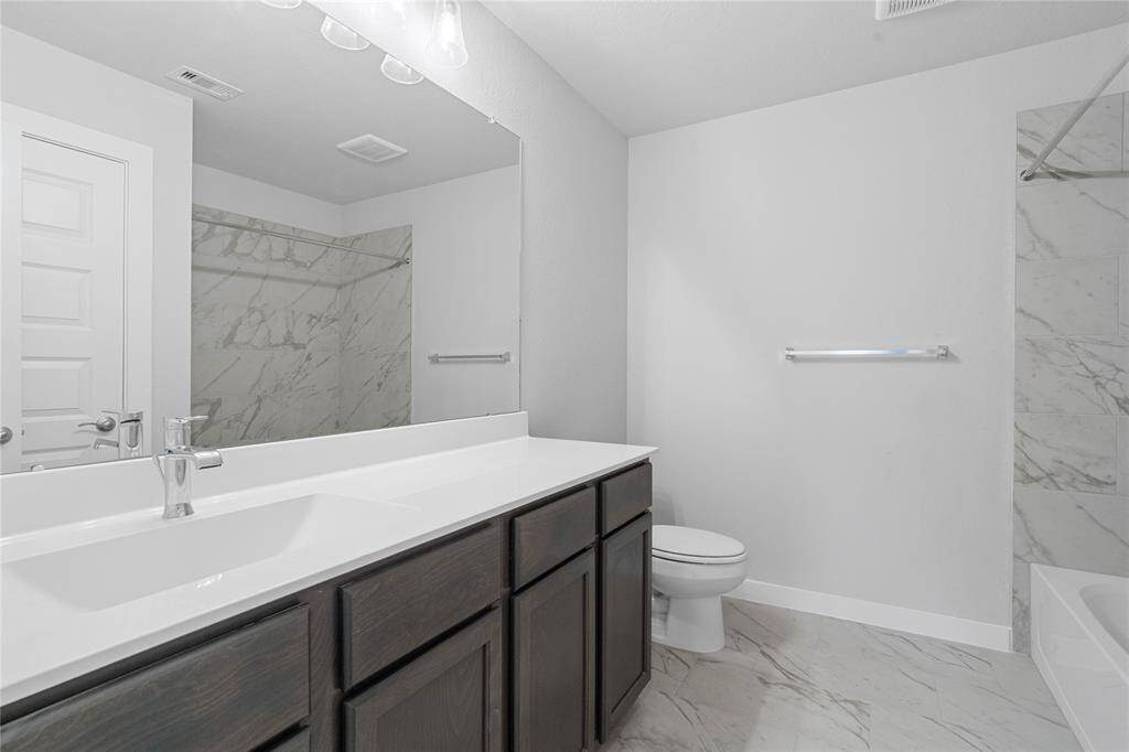 Secondary bath features tile flooring, bath/shower combo with tile surround, stained wood cabinets, beautiful light countertops, mirror, dark, sleek fixtures and modern finishes.