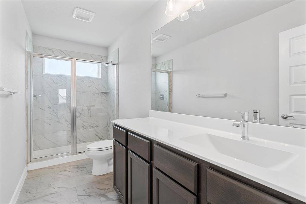 Secondary bath features tile flooring, large walk-in shower with tile surround, stained wood cabinets, beautiful light countertops, mirror, dark, sleek fixtures and modern finishes.
