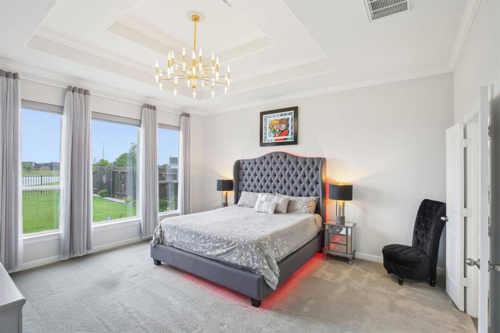 This spacious bedroom features large windows with elegant drapes, a stylish tray ceiling with a modern chandelier. Natural light fills the room, highlighting the soft carpet and neutral color palette for a serene, welcoming atmosphere.
