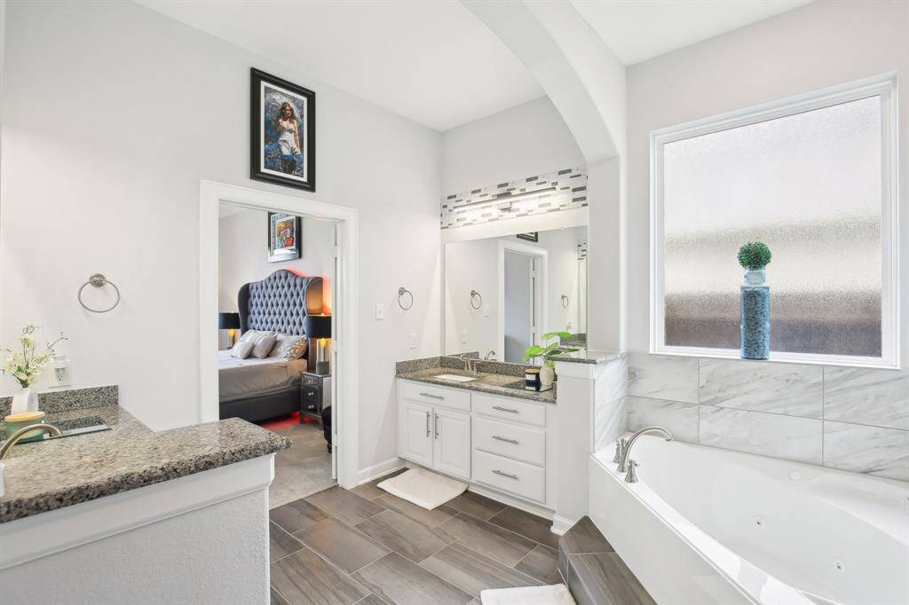 This bright bathroom features a luxurious soaking tub, sleek granite countertops, and modern cabinetry. The space is complemented by a large frosted window, elegant tile flooring, and an adjacent view into the stylish primary suite.