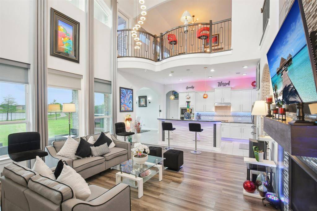 Check out this spacious, modern living area with high ceilings and large windows offering plenty of natural light. The open floor plan includes a stylish kitchen with a breakfast bar and sleek decor. A second-floor balcony overlooks the den, adding a touch of elegance.