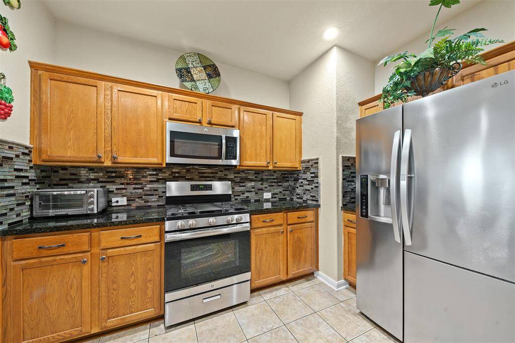 This kitchen features warm wooden cabinetry, modern stainless steel appliances, and a stylish tile backsplash. The space is enhanced by a sleek refrigerator and abundant overhead lighting, creating a bright and inviting cooking area.