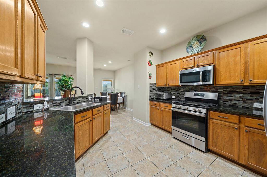 This kitchen features modern stainless steel appliances, wooden cabinetry, and a sleek black granite countertop. The open layout flows into a dining area, and the space is well-lit with recessed lighting.