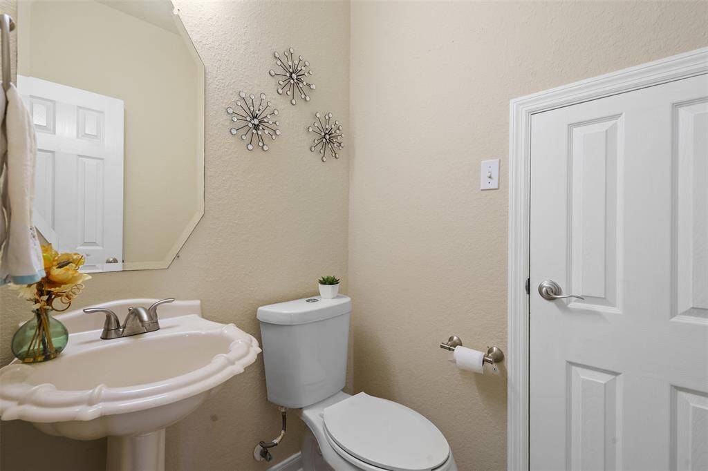 This is a compact, well-lit half-bathroom featuring a pedestal sink, toilet, decorative wall accents, and a small mirror. It has neutral-colored walls and a simple, clean design.