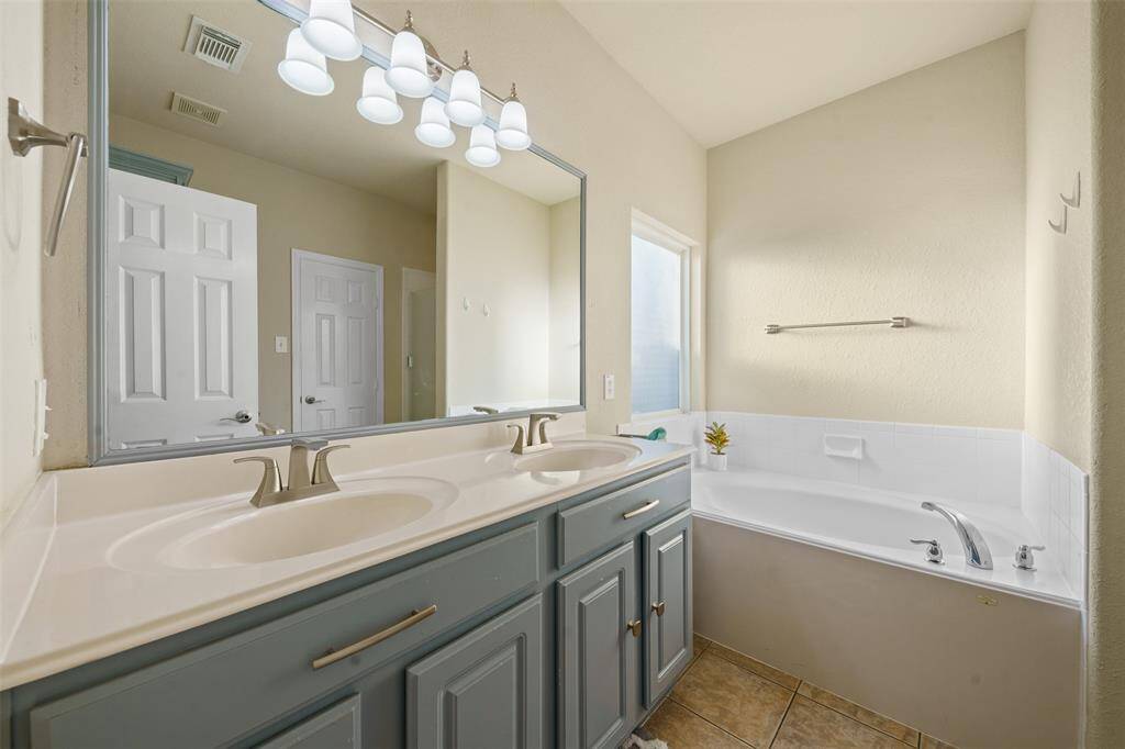 This bathroom features a double sink vanity with ample storage, a large mirror, modern lighting, and a spacious soaking tub, all in a bright, neutral-toned space.