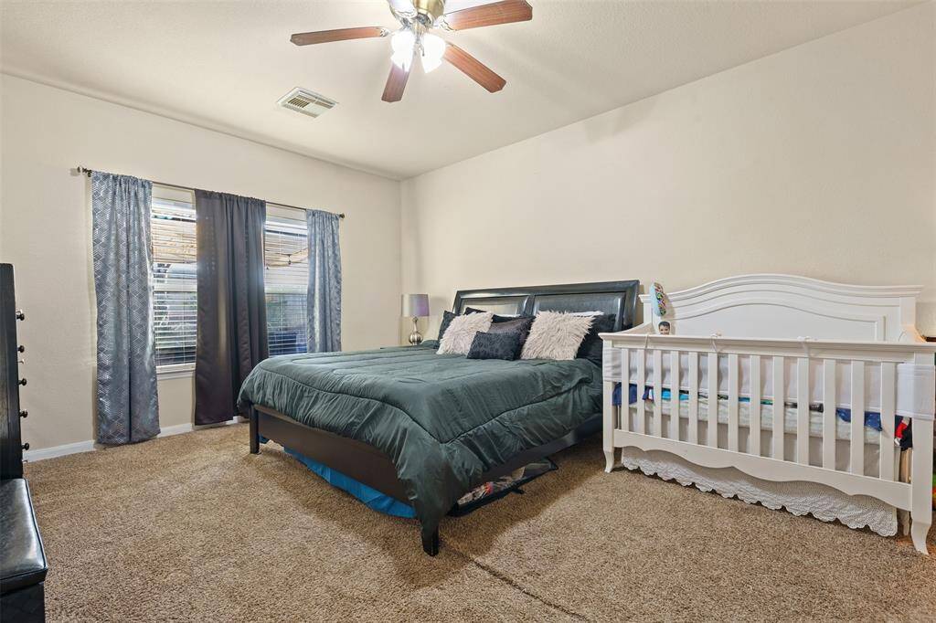 This spacious bedroom features a comfortable layout with a large bed, a crib, and neutral carpeting. Natural light streams through the window, complemented by elegant curtains. A ceiling fan provides additional comfort.