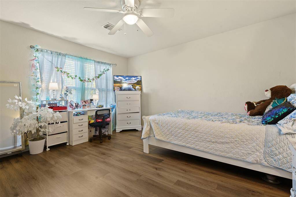 This bright bedroom features wood flooring, a ceiling fan, and neutral walls. It includes a single bed, a desk with decorative touches, and a dresser, creating a cozy and functional space.