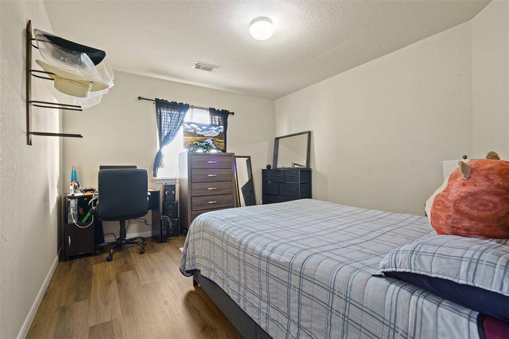 This cozy bedroom features a comfortable bed, a small desk with a chair, and ample storage with dressers. The room is well-lit with natural light from the window, complemented by soft overhead lighting. The hardwood floors add warmth to the space.