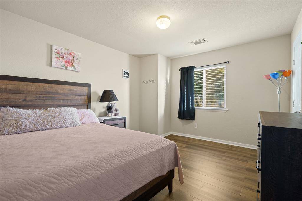 Cozy bedroom with a wooden floor, featuring a bed with a stylish headboard, soft bedding, and a bedside table with a lamp. The room has natural light from a window with dark curtains, complemented by neutral wall colors and simple decor.