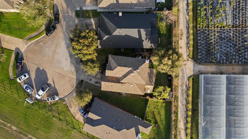 This aerial photo shows a residential area with several houses arranged around a cul-de-sac. The properties are surrounded by green lawns and some trees. Adjacent to the homes, there's an agricultural field or nursery with rows of plants. The setting appears suburban and tranquil.