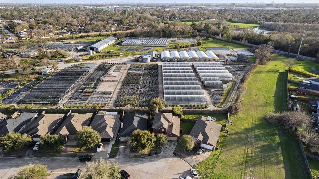 This aerial photo showcases a residential neighborhood with neatly arranged houses and a nearby large greenhouse complex surrounded by greenery. Ideal for someone seeking a home close to agricultural or gardening facilities.