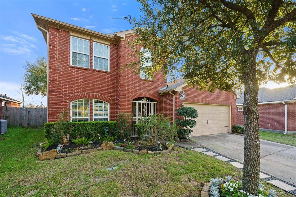 This is a two-story brick home with a well-maintained front yard featuring a tree, shrubs, and stone-bordered flower beds. It has a two-car garage and a gated entrance, offering a welcoming and secure exterior.