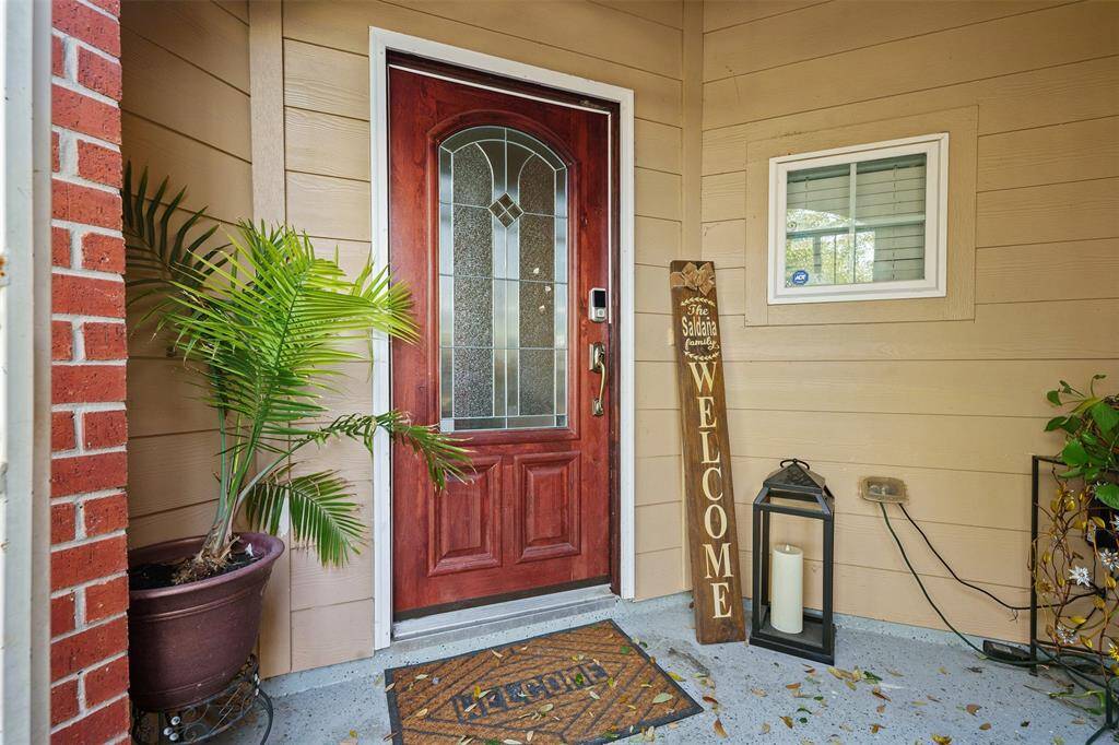 This welcoming front entrance features a rich wooden door with decorative glass panels, a potted plant, and a charming welcome sign. The area is accented with a small window and a lantern, creating a cozy and inviting atmosphere.