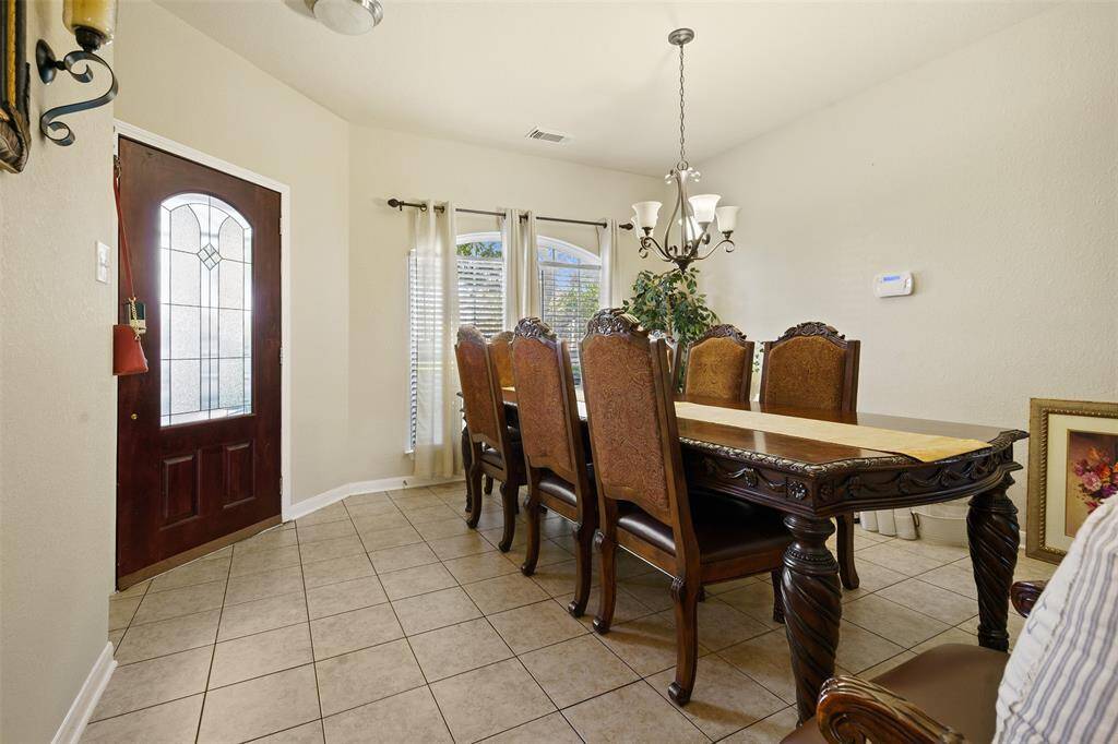 This photo features a dining area with a large wooden table and eight ornate chairs. The room has tiled flooring, a chandelier, and natural light from a window with blinds. A decorative door and neutral wall colors create a welcoming atmosphere.