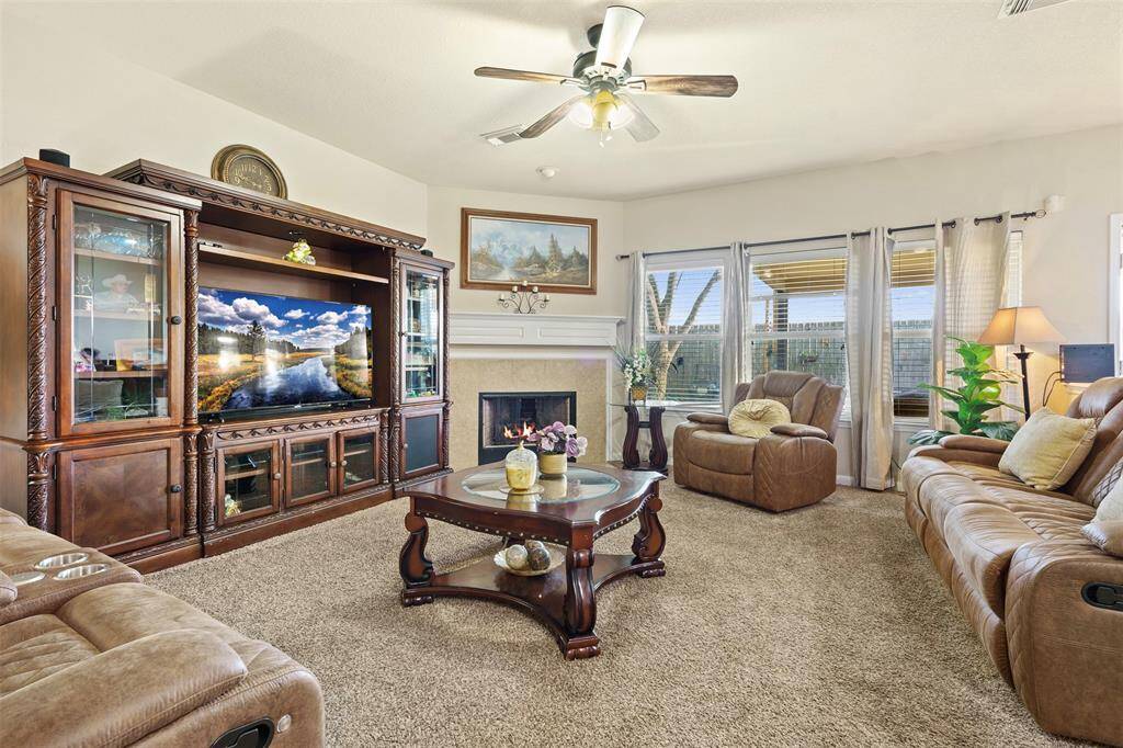 This cozy living room features plush carpet, a large entertainment center, and comfortable leather seating. It includes a fireplace, ceiling fan, and large windows with natural light, offering a warm and inviting atmosphere.