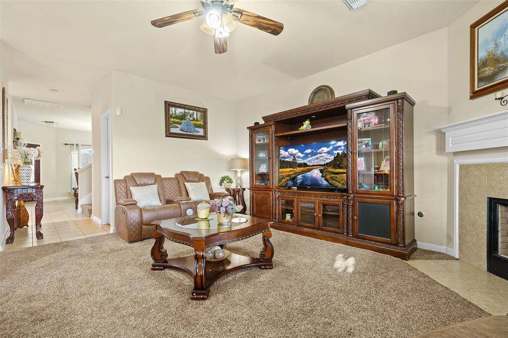 This cozy living room features a spacious layout with a plush carpet, elegant wooden furniture, and a large entertainment center. It includes a comfortable seating area, a ceiling fan, and a fireplace, creating a warm and inviting atmosphere.