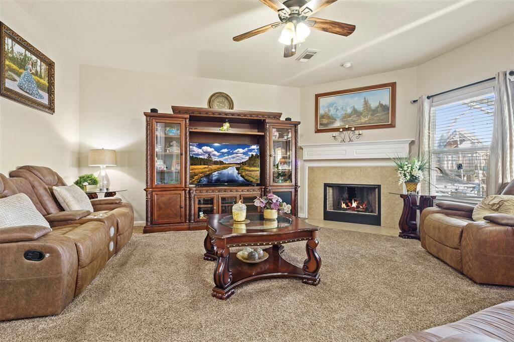 This cozy living room features plush carpeting, a warm fireplace, and comfortable leather seating. It includes a large wooden entertainment center with built-in shelving and a TV. Natural light fills the space through a large window, enhancing the inviting atmosphere.