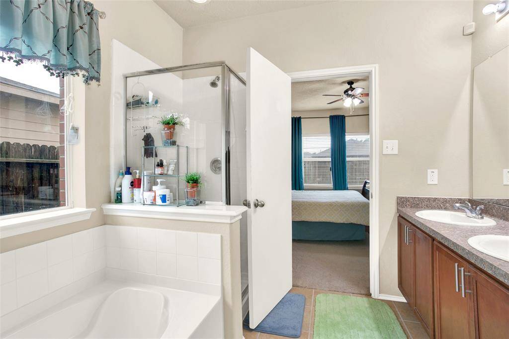 Experience the harmony of design and functionality in this bright, attached master bath featuring a double vanity, expansive mirror, soaking tub, and separate shower. Admire the seamless flow into the tranquil bedroom, complete with a cooling ceiling fan, visible beyond the bathroom's entrance.