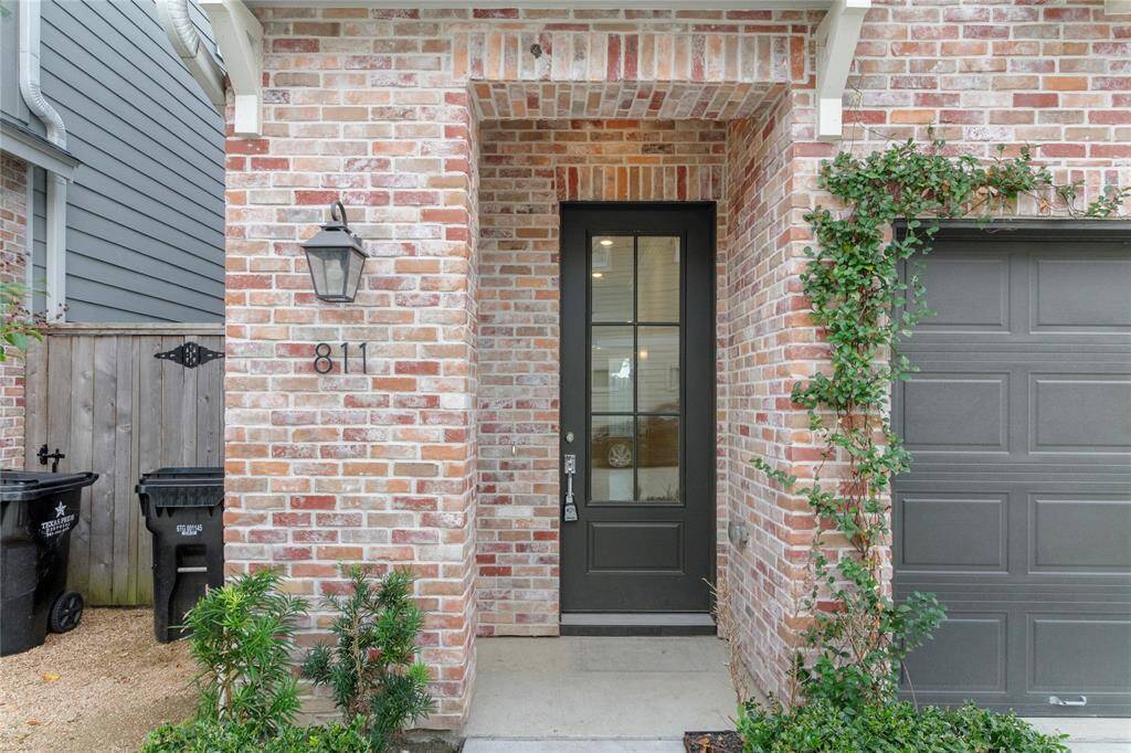 Charming brick exterior with a welcoming front entry, featuring a glass-panel door and a classic lantern-style light fixture. There’s a garage to the right and a small garden area with greenery adding a touch of nature.